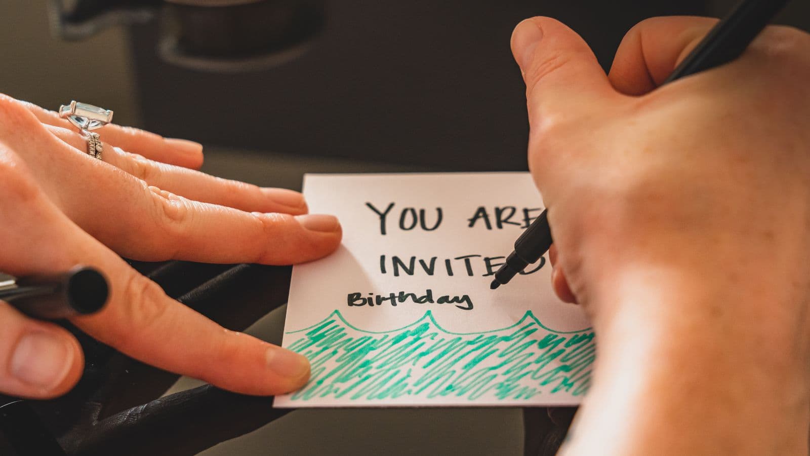 A close-up of hands carefully hand-lettering a colorful "You Are Invited" birthday card.