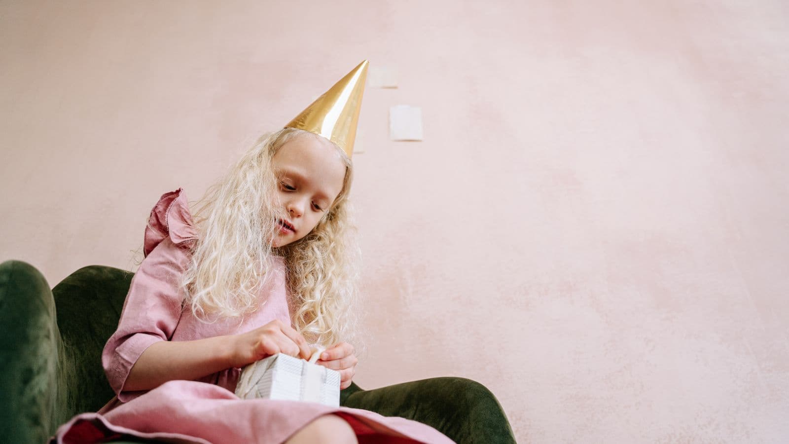 A young girl in a gold party hat sits in a green armchair, carefully untying the ribbon on a white wrapped gift.