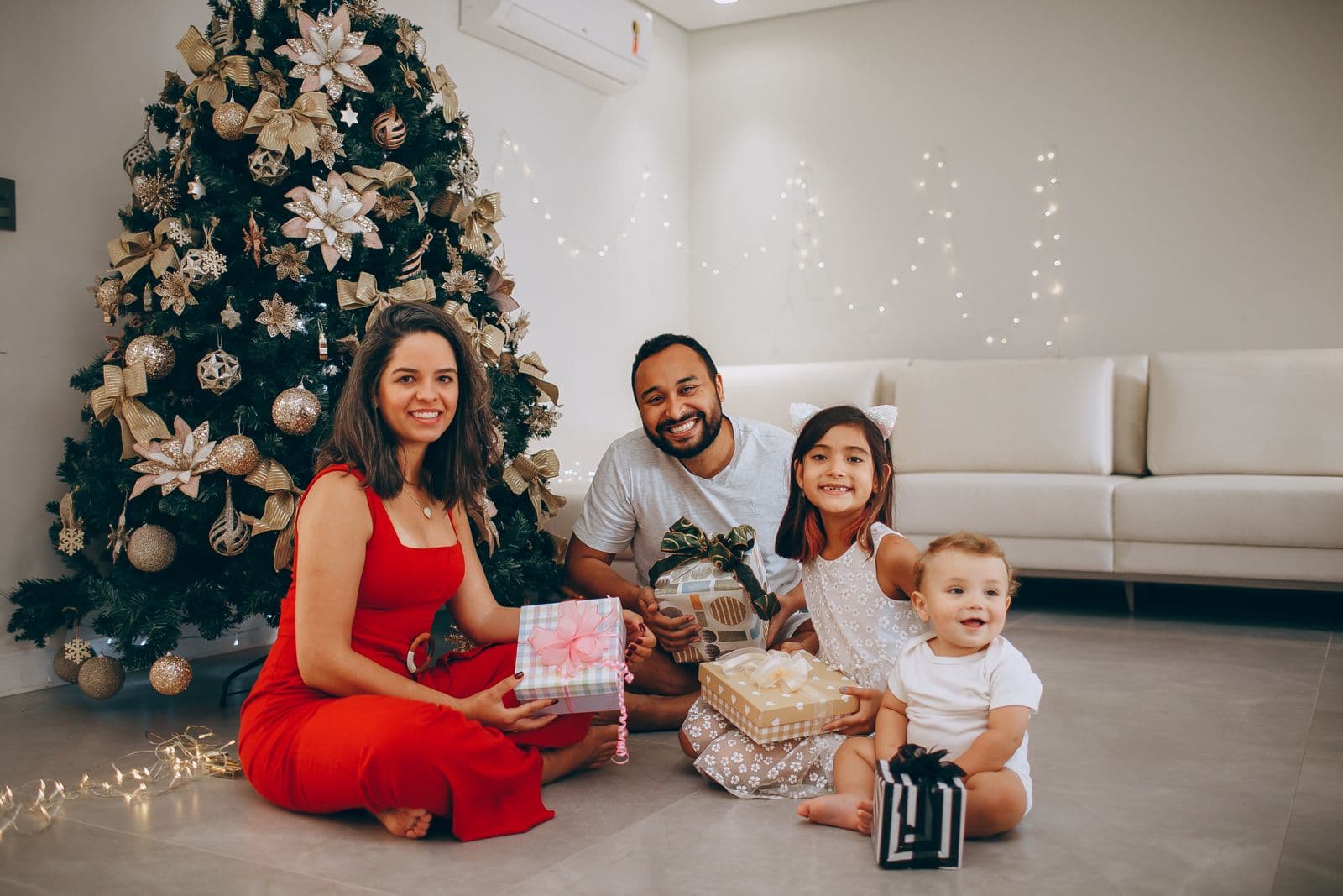 Parents and two young kids holding wrapped gifts beside the Christmas tree.