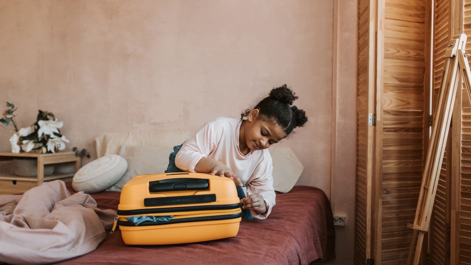 A smiling child closing a bright yellow suitcase on a bed in a warm, pastel-toned bedroom.