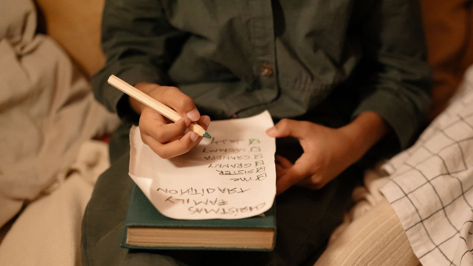 A child's hand writing a family gift checklist by pencil in warm, cozy lamplight.