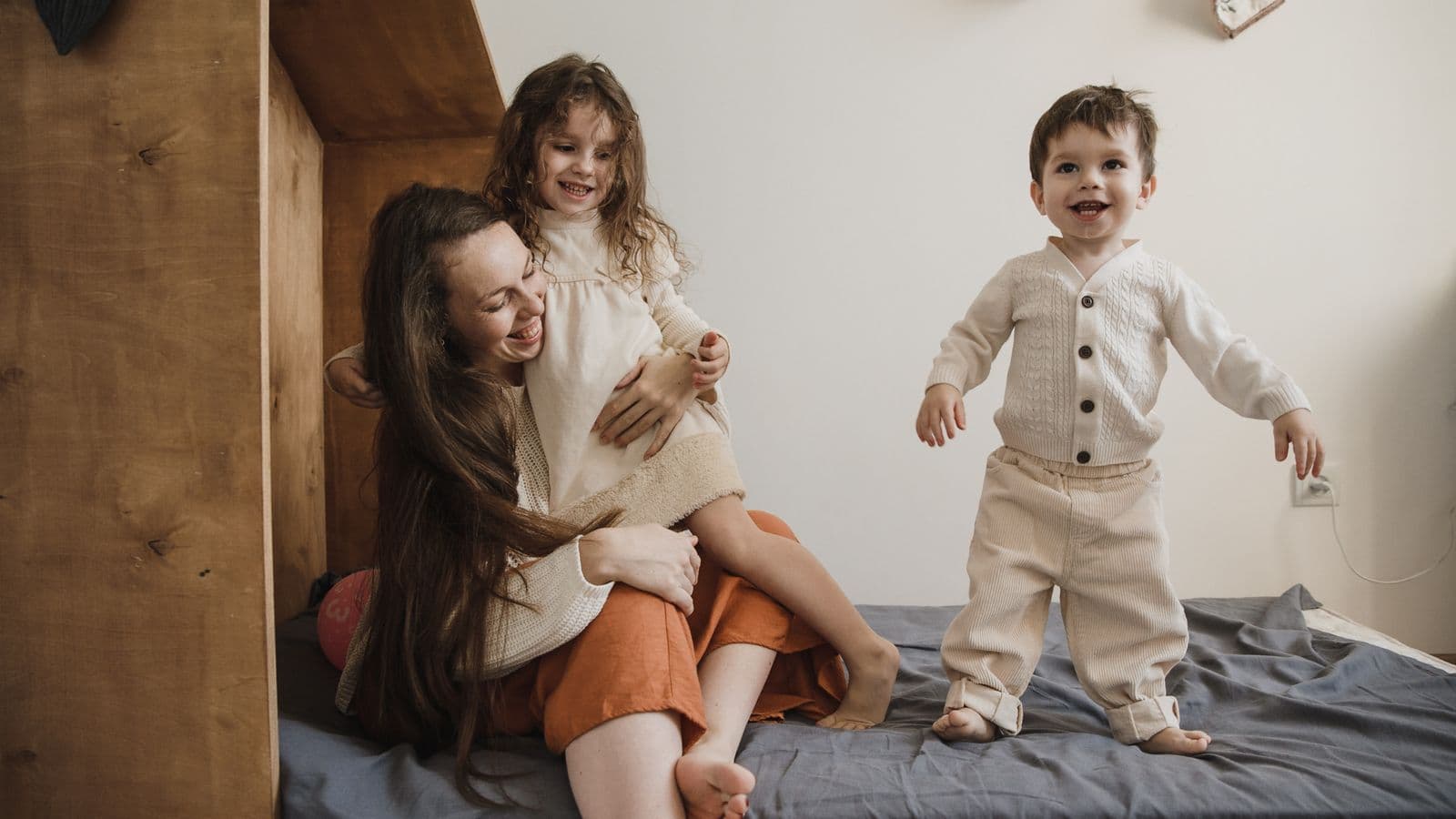 A mother laughing with her two young children, one held close and one standing, in a warm cream-toned bedroom.
