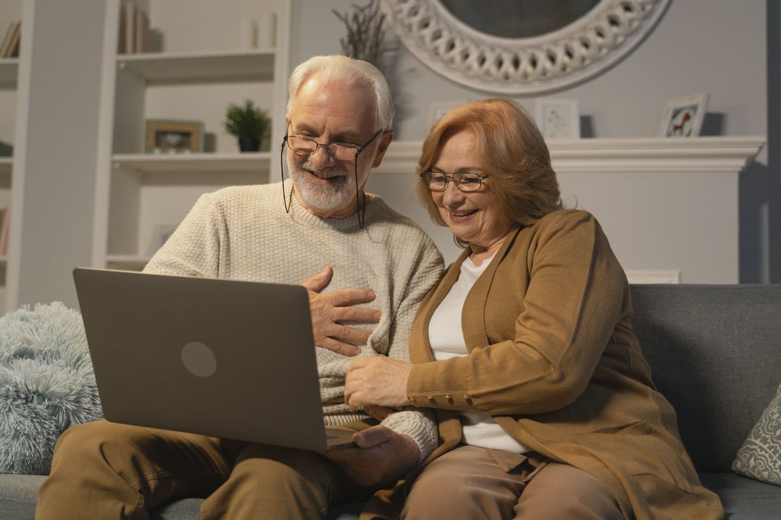 A smiling grandparent couple on a couch sharing a laptop video call.
