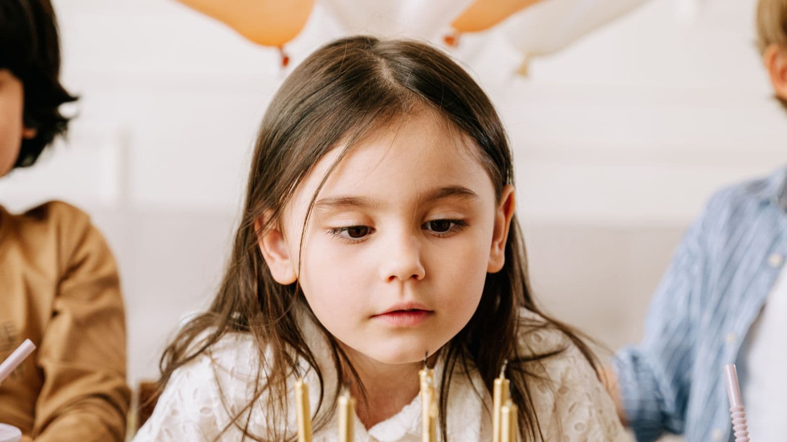 A young girl gazes at the glowing candles on her birthday cake while balloons float softly in the background.