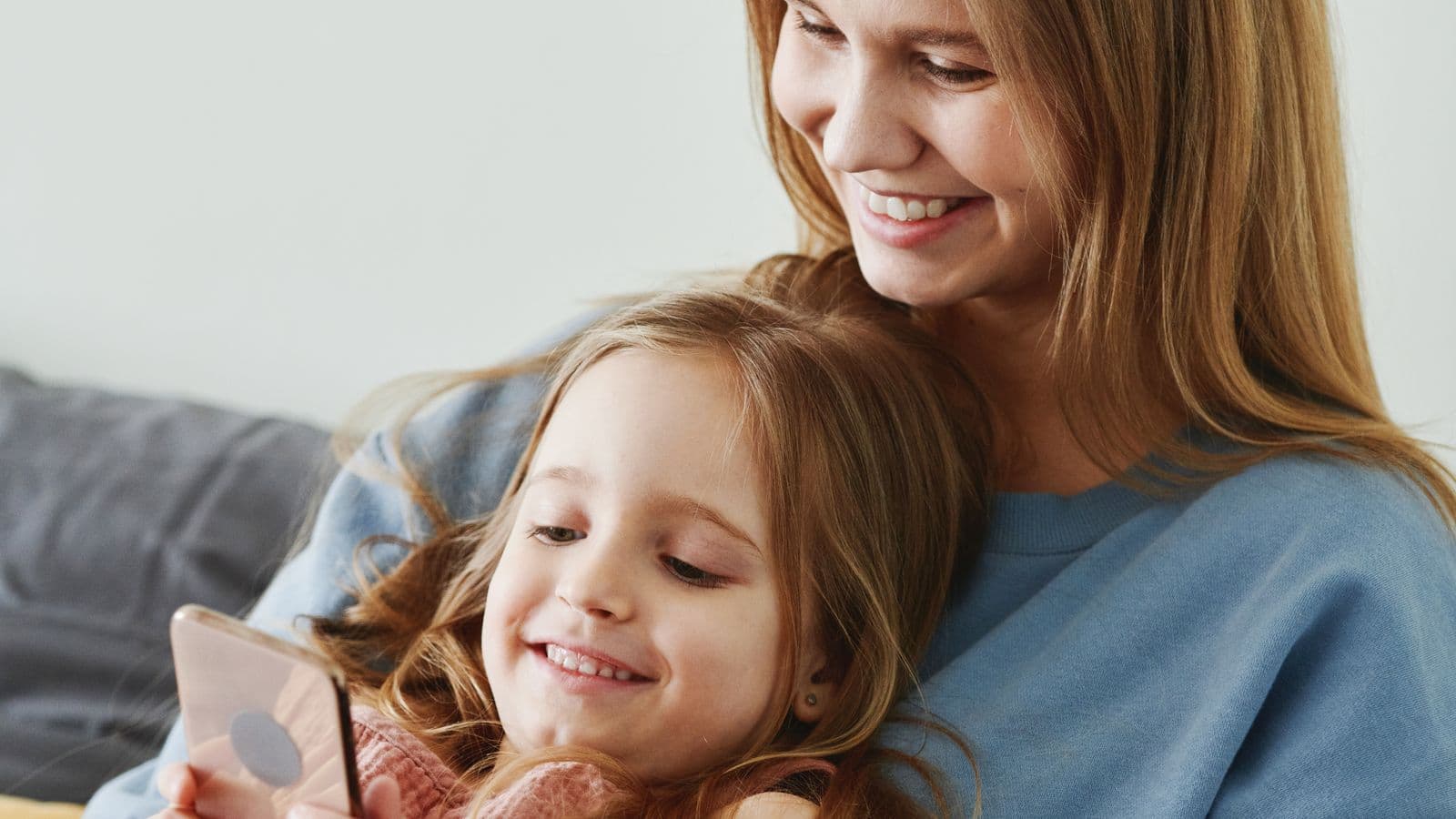 A mother in a soft blue sweater and her young daughter sharing a smile while looking at a smartphone together at home.
