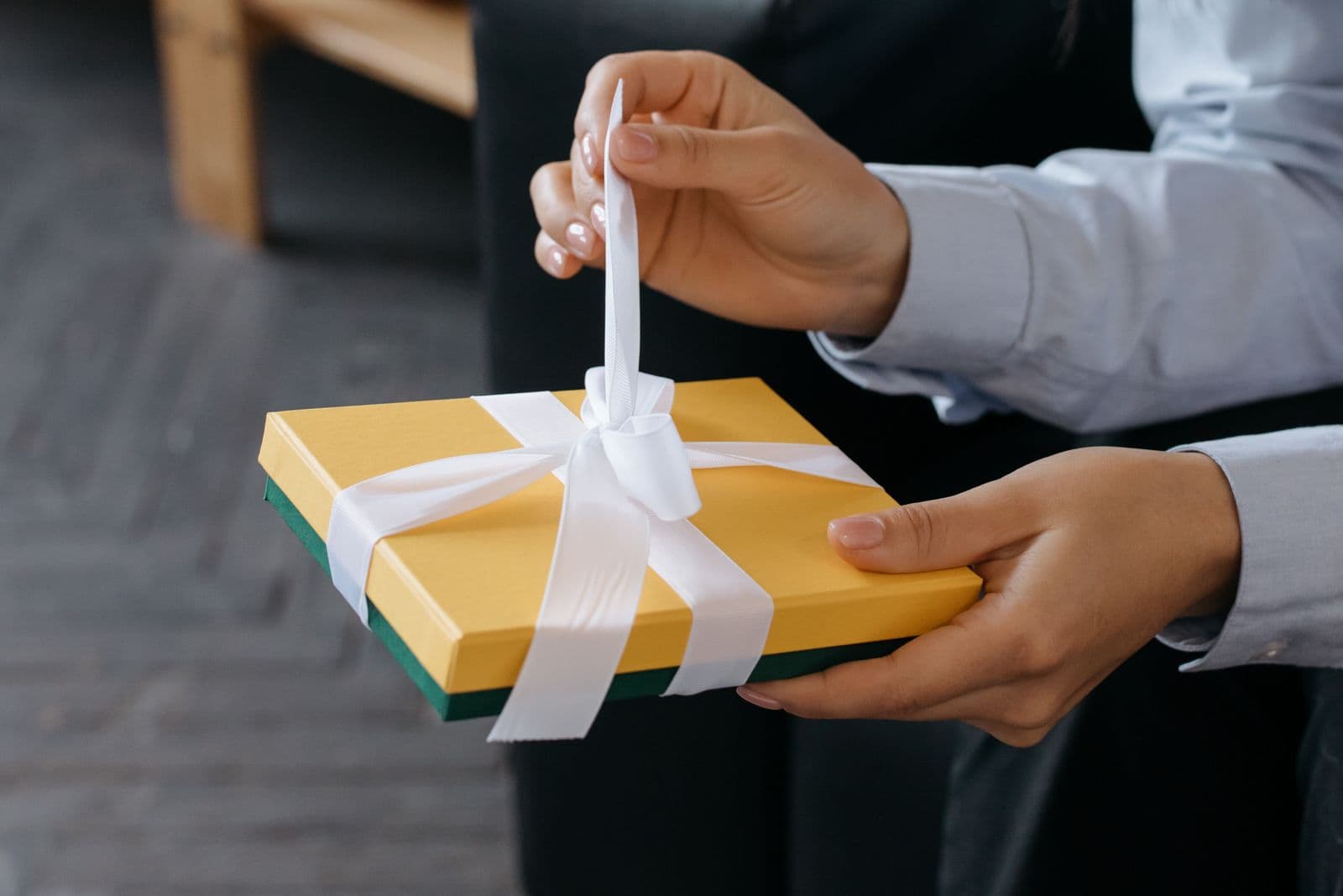 Hands untying the white ribbon bow on a yellow gift box.
