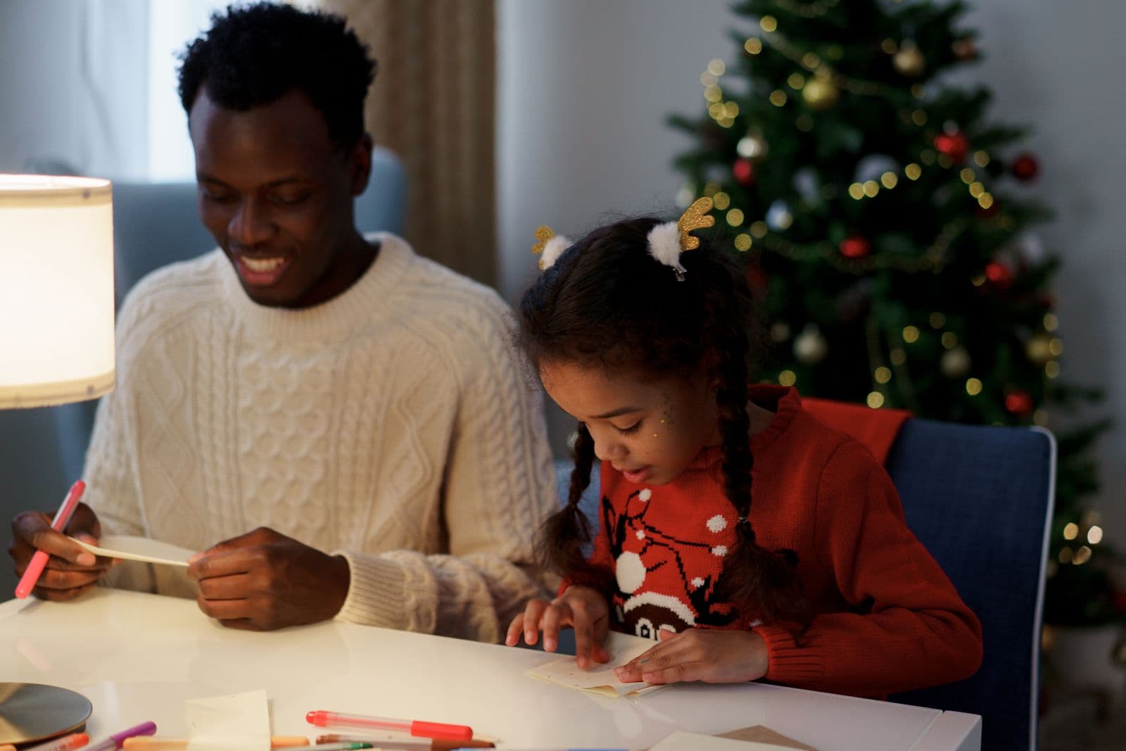 A dad and his daughter writing a Christmas list together at a table beside the tree.