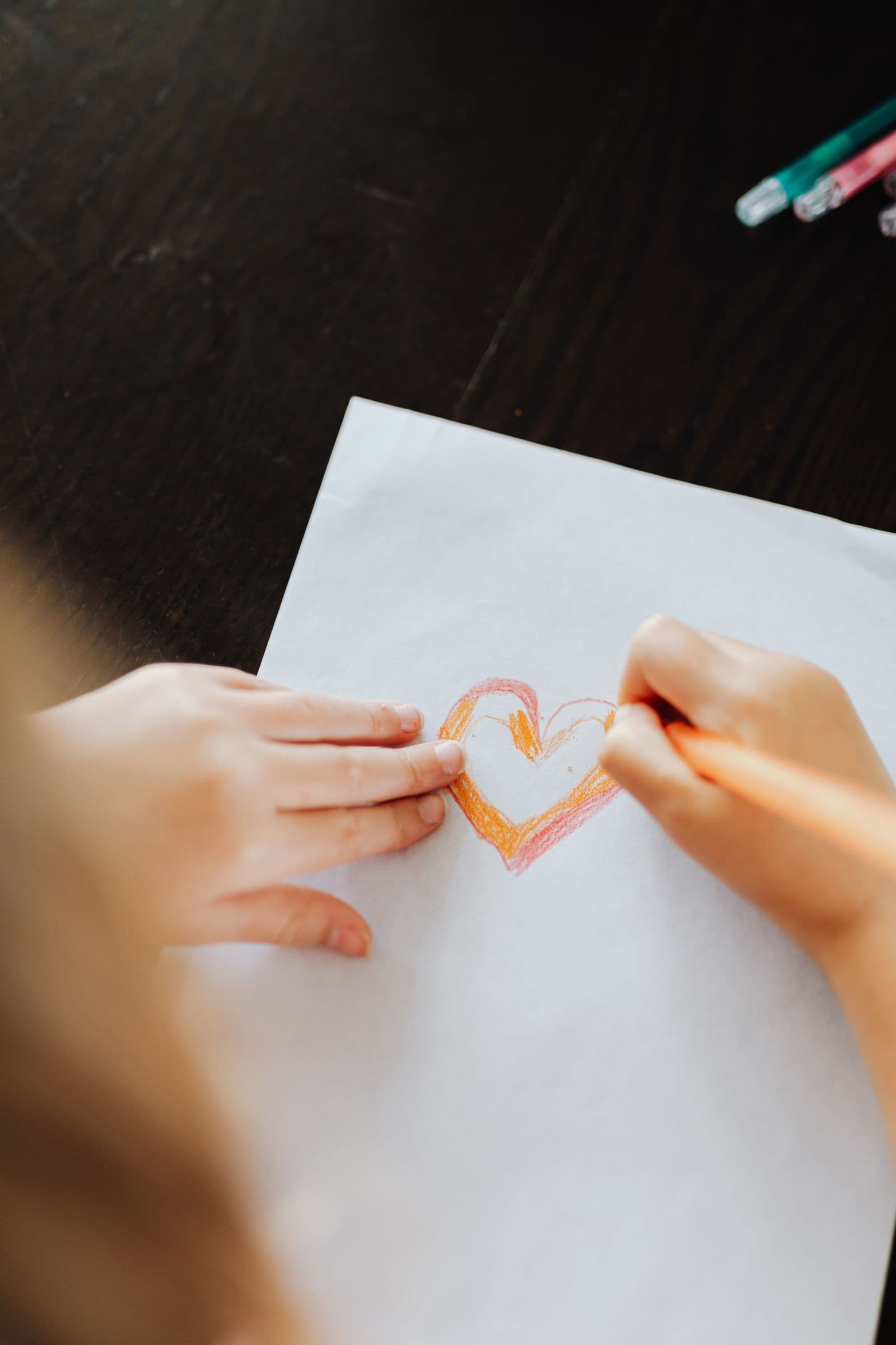 A child’s hands drawing an orange heart on a homemade card at a wooden table.